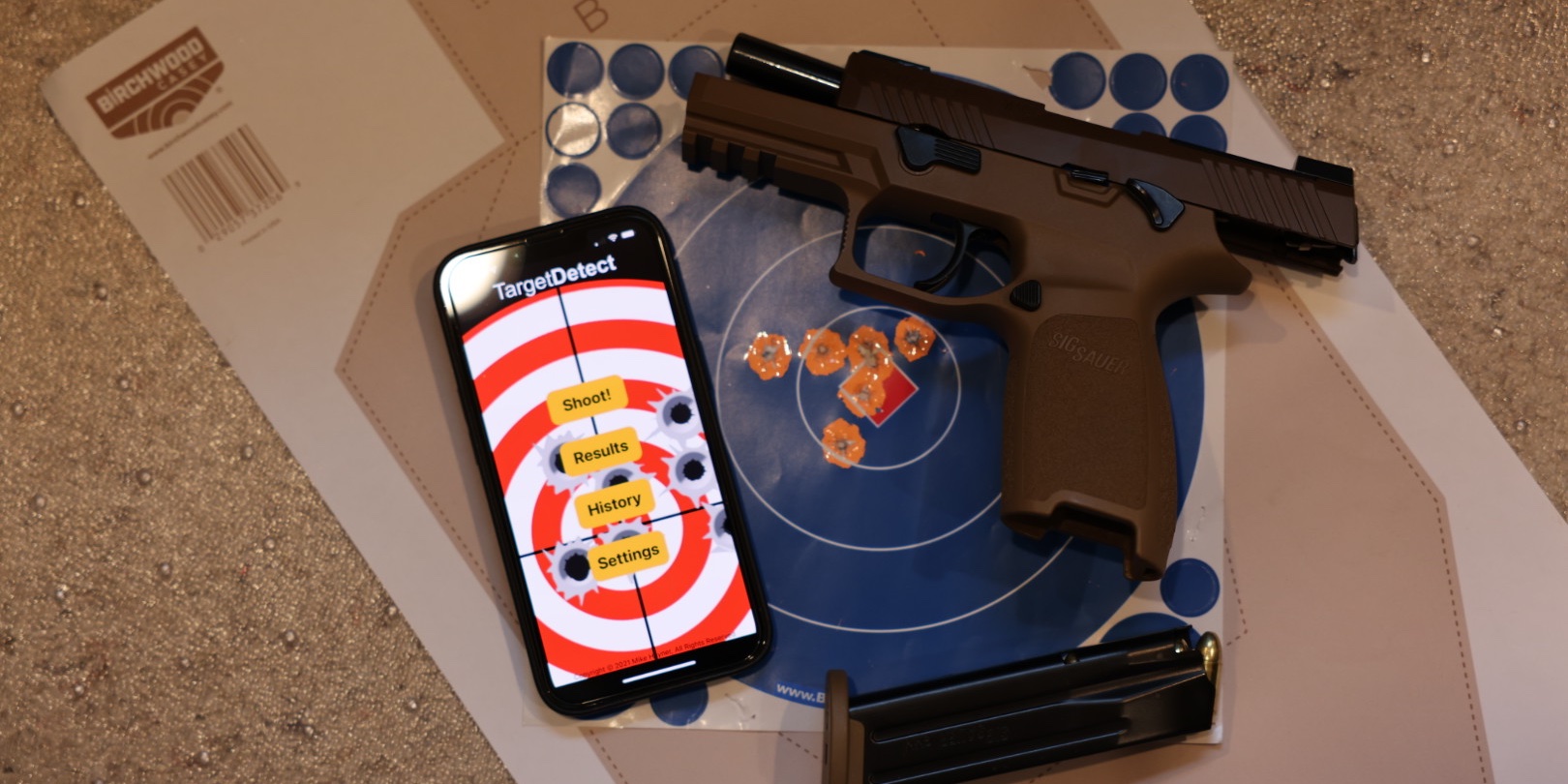 Target Detect on iPhone, lying on a Birchwood Casey bullseye target alongside a Sig Sauer pistol and magazine — a scorebook for the modern sporting shooter